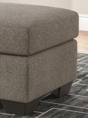 Ashley Furniture Triston-Peaks Ottoman showing a wooden frame with fabric upholstery and decorative details.