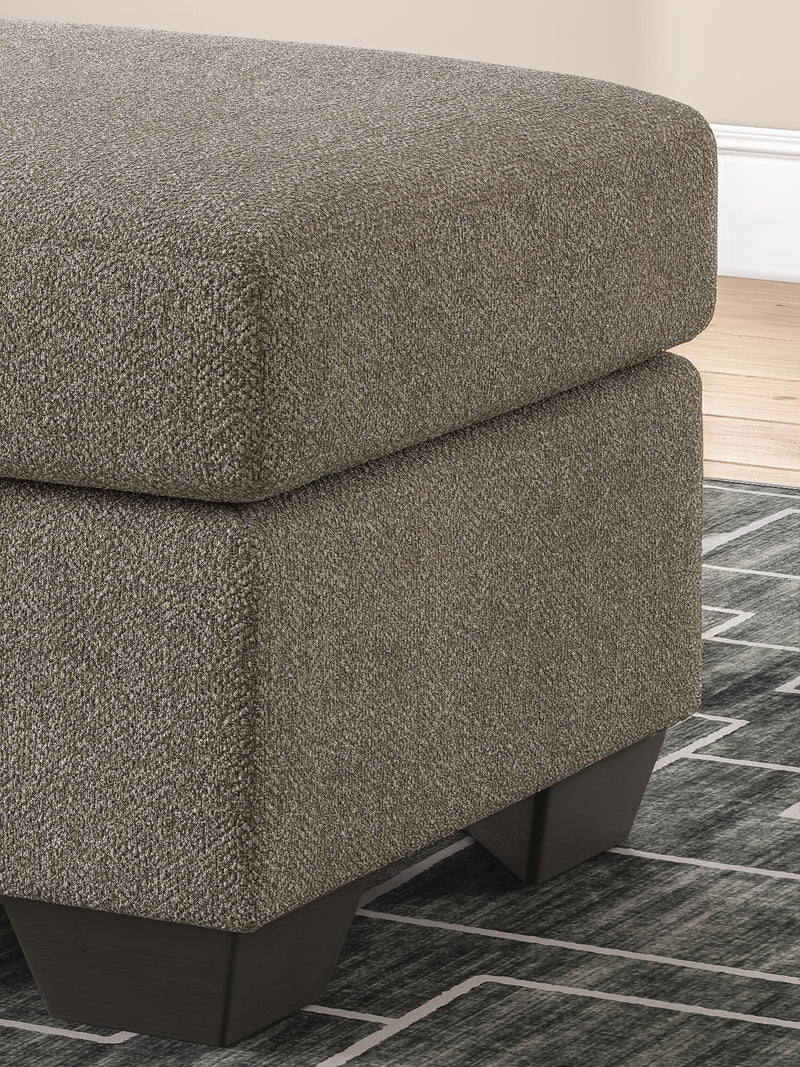 Ashley Furniture Triston-Peaks Ottoman showing a wooden frame with fabric upholstery and decorative details.