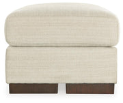 Ashley Furniture Maggie Ottoman with soft fabric upholstery and wooden legs, shown in a cozy living room setting.