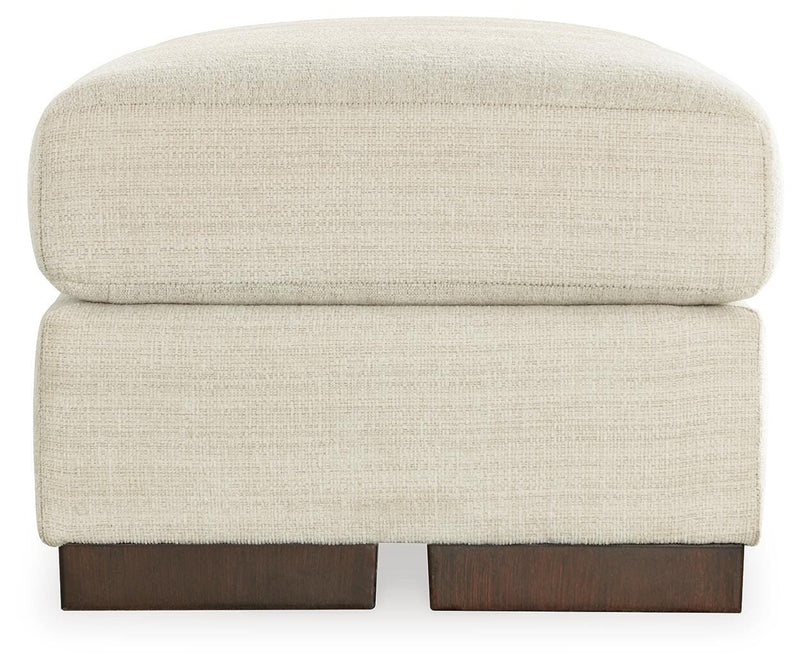 Ashley Furniture Maggie Ottoman with soft fabric upholstery and wooden legs, shown in a cozy living room setting.
