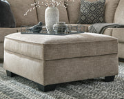 Ashley Furniture Bovarian Ottoman in beige fabric with tufted top and wooden legs, resting on a hardwood floor.