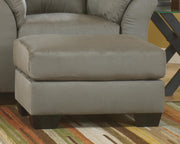 Ashley Furniture Darcy Ottoman in beige fabric with tufted top and wooden block legs, ideal as a versatile living room accent piece.