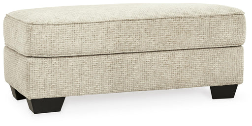 Ashley Furniture Monaghan Ottoman with plush fabric, cushioned top, and wooden legs, shown from an angled perspective.