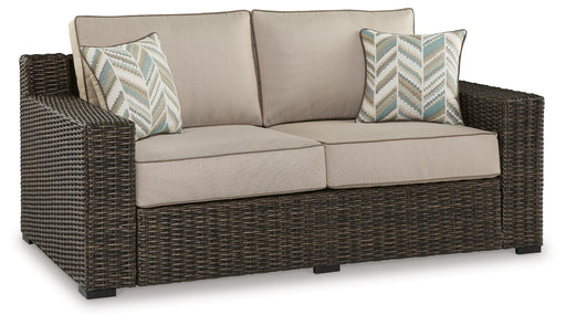 Ashley Furniture Coastline Bay outdoor loveseat with cushion, featuring woven frame and comfortable seating, shown from an angled view.